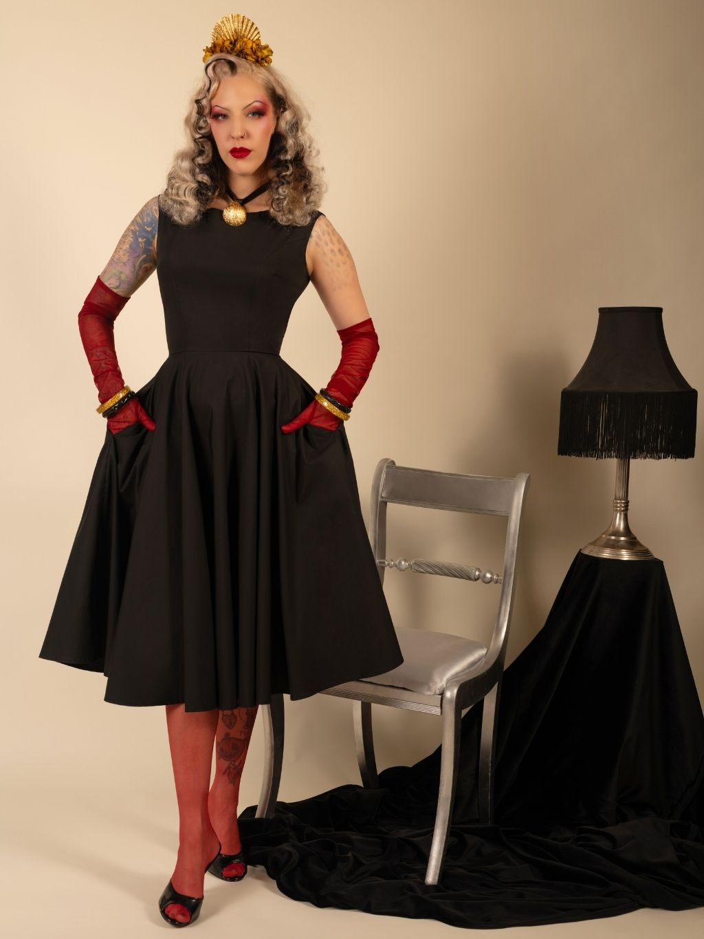 Woman in a black dress with red gloves and shoes posing next to a chair and lamp.