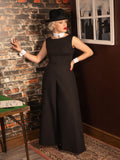 Woman in vintage black beach pyjamas and hat standing against a brick wall.