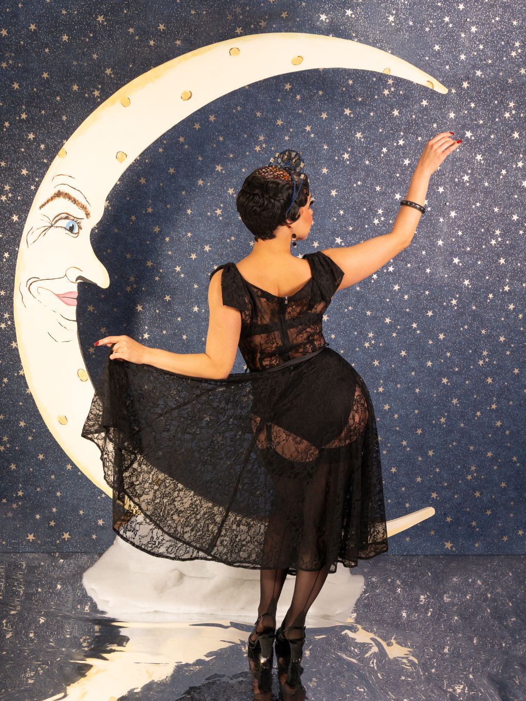 Woman in a black dress standing next to a crescent moon with a face on a starry background