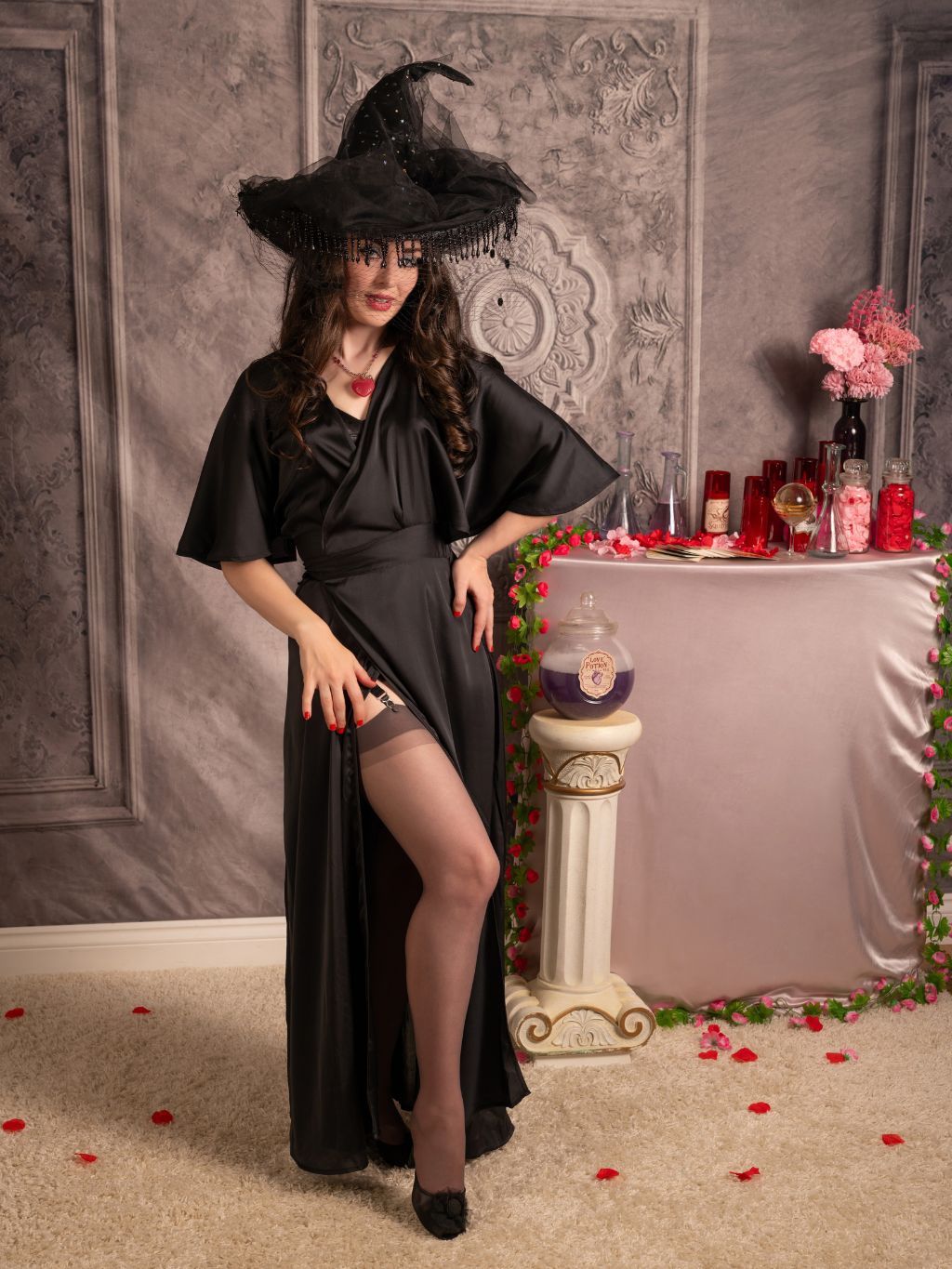 A woman models a black witch costume dress with a matching pointed hat, posing in a themed room with a pink table, rose petals, and gothic wall decor.