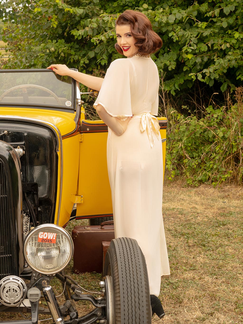Woman in a peach satin robe next to a vintage yellow car with greenery in the background