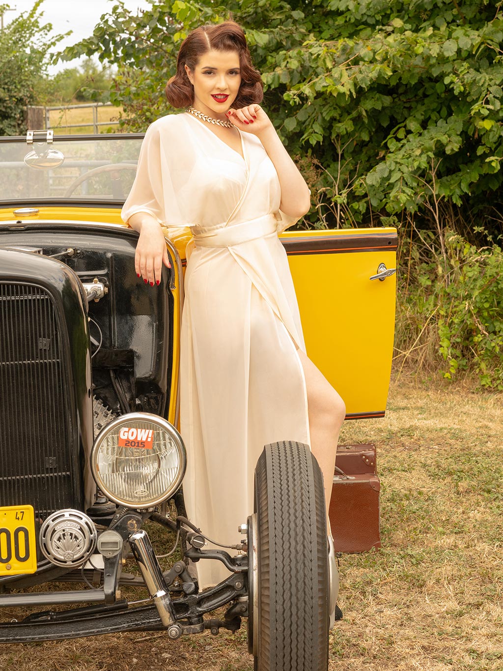 Woman in a long peach satin robe standing next to a vintage yellow car with greenery in the background